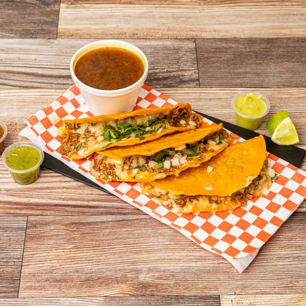 Three Chonchis birria tacos with consommé, green salsa, and lime wedges on a wooden table.