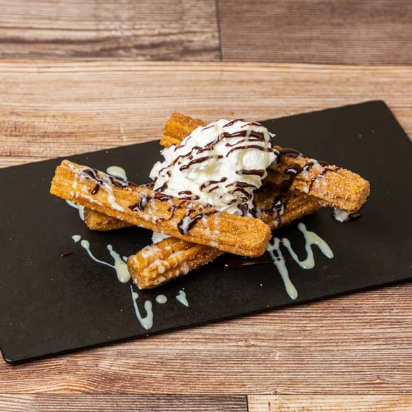 Churros with whipped cream, chocolate, and white sauce drizzle on a black plate.